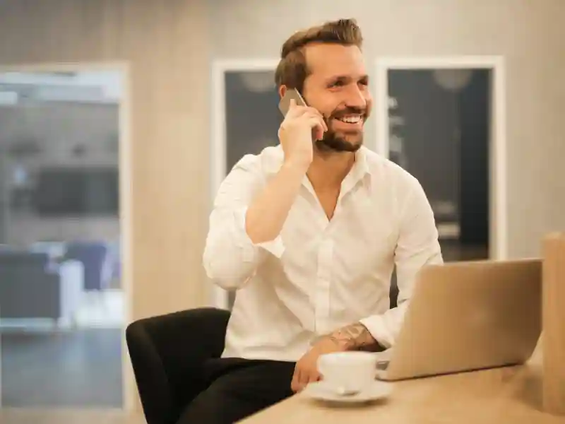 A man in a white button-down shirt smiles while talking on the phone in an office, with a laptop and cup in front of him